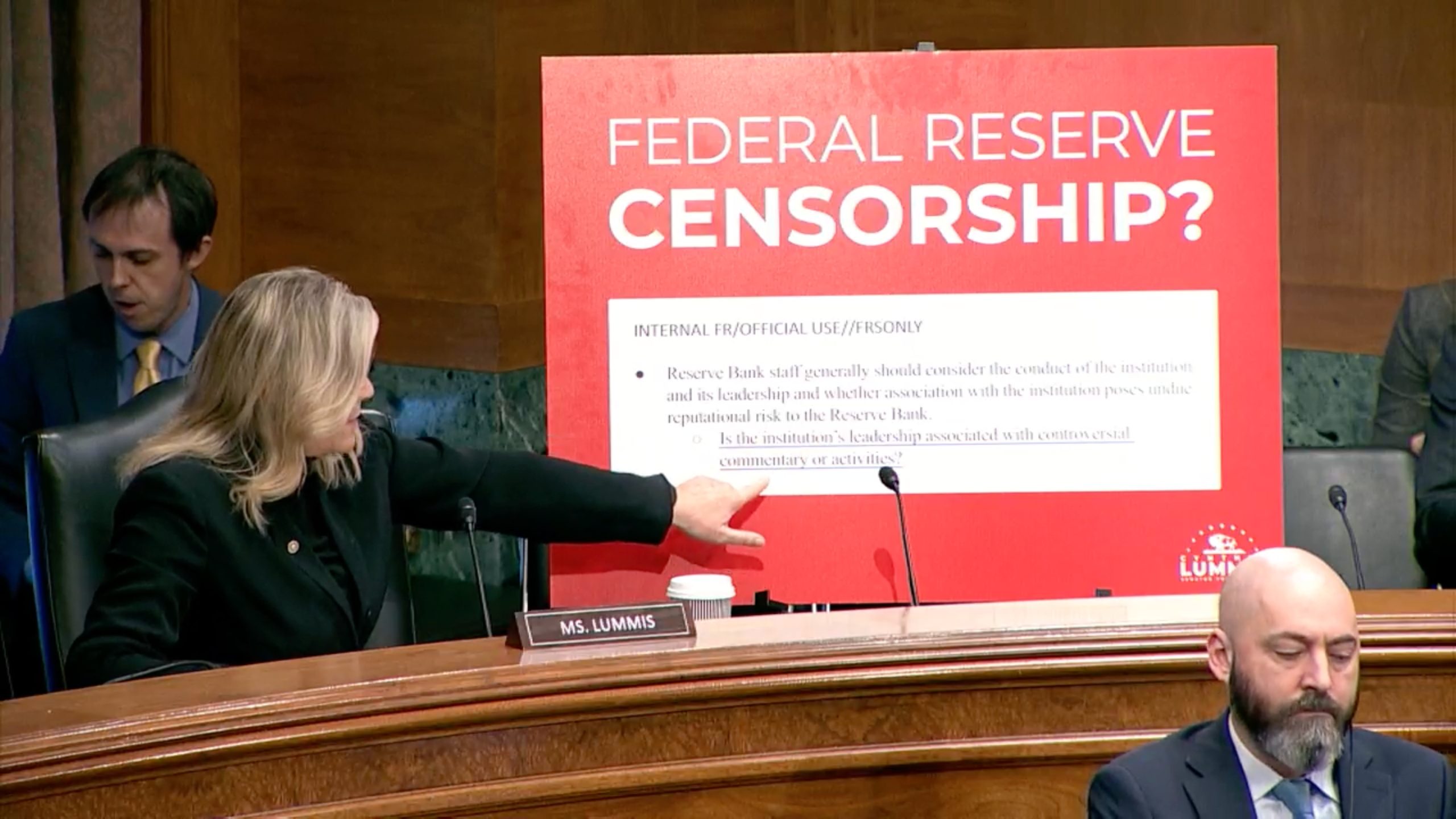 Senator Cynthia Lummis (R-WY) sits at a desk with a microphone, next to a placard labeled "Ms. Lummis" while pointing at a large document in the background which contains a note surrounded by a red border. The document shows a quote from a confidential Federal Reserve implementation handbook on account actions that requires Federal Reserve staff to "consider the conduct of the institution and its leadership and whether association with the institution poses reputational risk to the Reserve Bank."