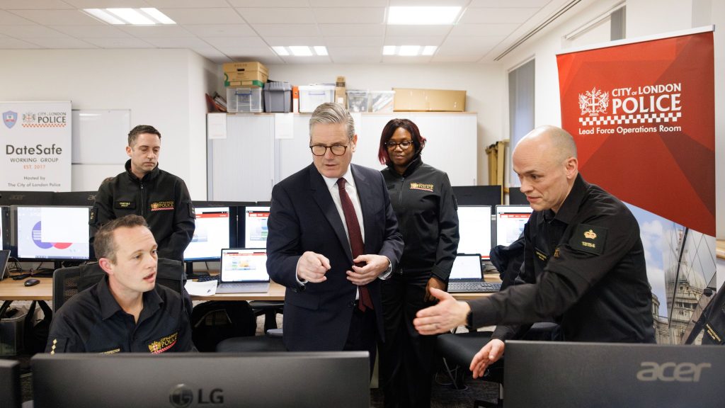 Starmer in a City of London Police operations room, engaged in discussion, with computers and a banner visible in the background.