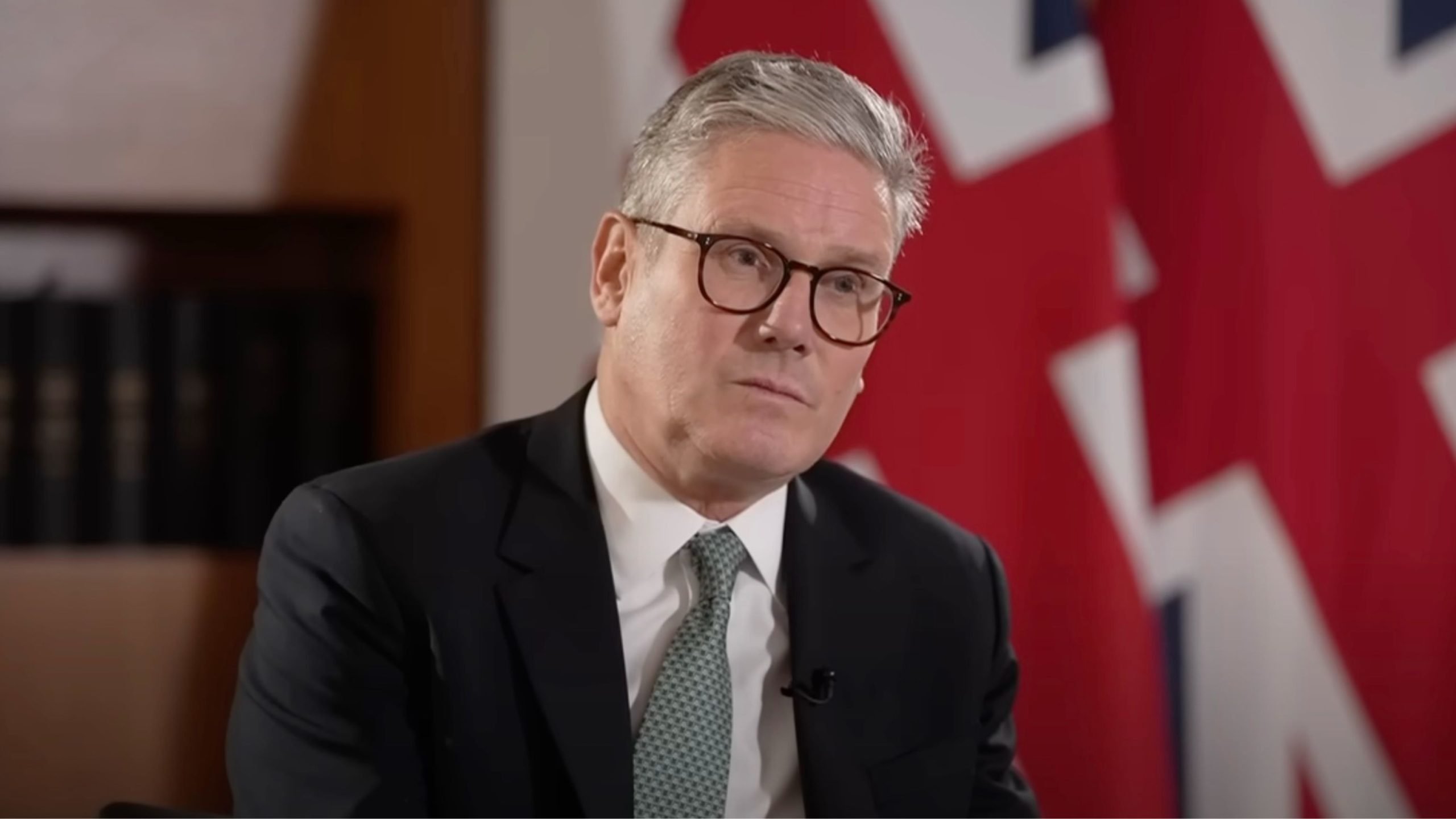 Starmer in a suit and glasses is sitting in front of a blurred Union Jack flag.