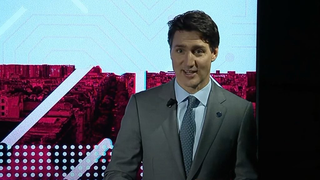 Trudeau in a suit speaking in front of a backdrop with red and white geometric patterns.