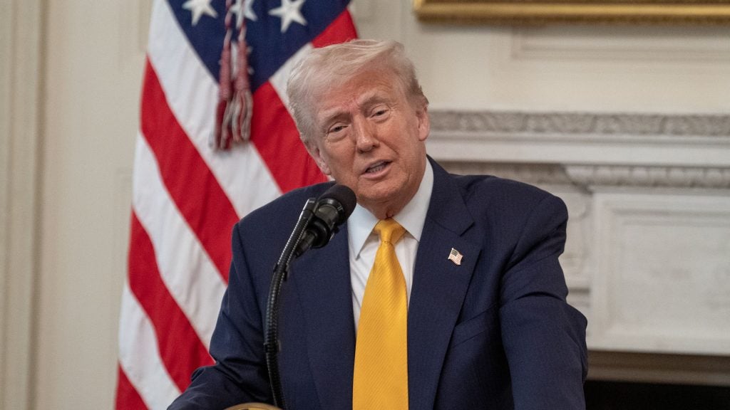 President Trump in a suit and yellow tie speaks at a podium with a microphone, with a U.S. flag in the background.