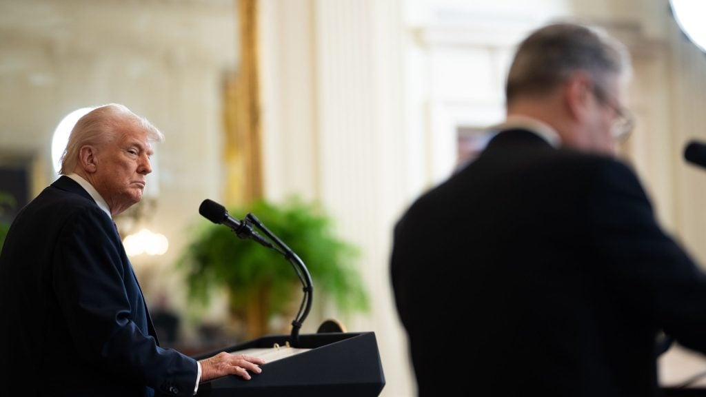 Trump and Starmer in a suit stands at a podium speaking into a microphone, with a blurred person in the foreground and a bright indoor setting with greenery in the background.