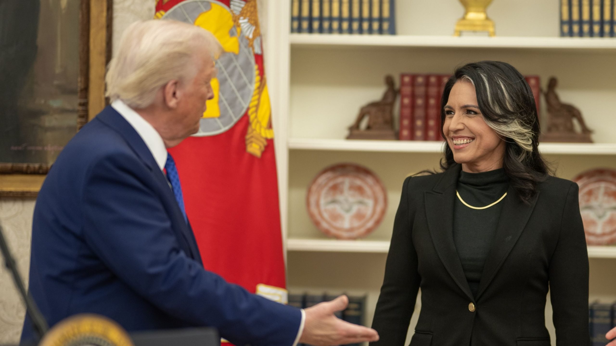 President Trump in a suit reaches out to Tulsi Gabbard in a blazer who is smiling in an office setting with bookshelves in the background.