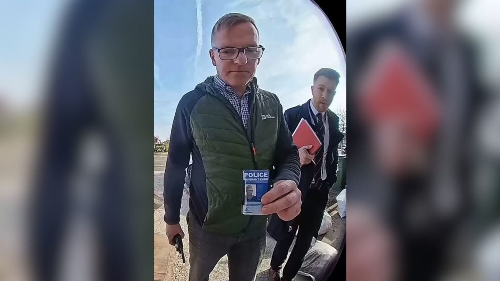 Two police men are standing at a doorway, one holding a police warrant card towards the camera. The man with the card wears glasses and a green jacket, while the other wears a suit and holds a red notebook.