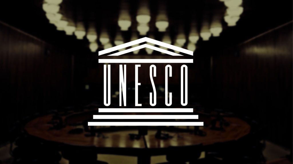 The UNESCO logo overlaying a dimly lit conference room with a circular wooden table and ceiling lights.