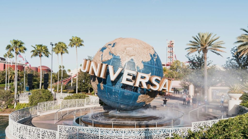 Globe statue with "UNIVERSAL" text at Universal Studios entrance, surrounded by palm trees and people walking nearby.