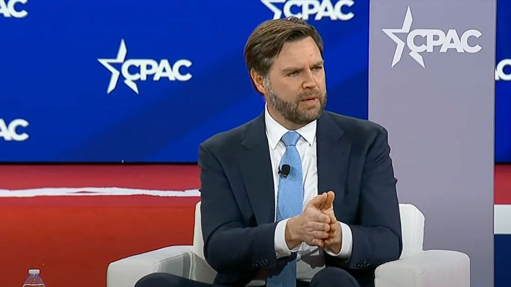 JD Vance in a suit and tie is speaking at a CPAC event, seated on a stage with a blue and red backdrop featuring the CPAC logo.