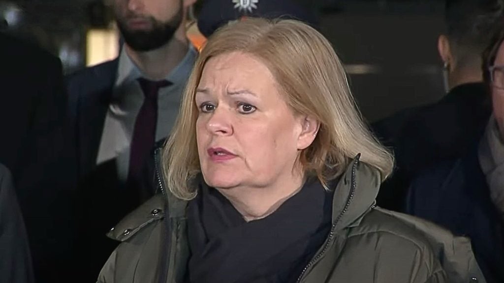 Faeser with blonde hair wearing a dark jacket and scarf speaks in front of a microphone, with several people visible in the blurred background.