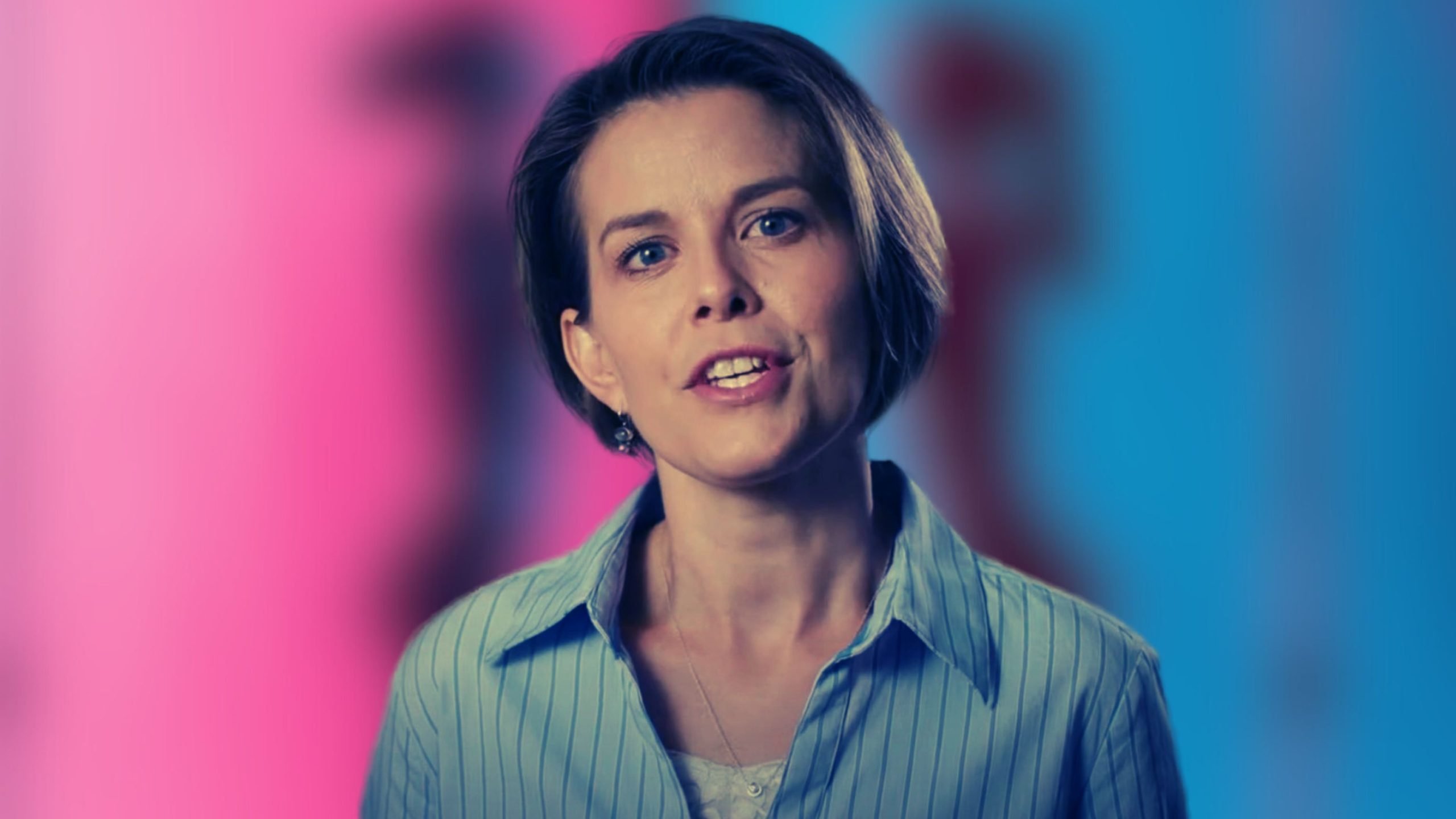 Laurel Libby with short brown hair and blue eyes, wearing a blue striped shirt, stands in front of a blurred pink and blue background.