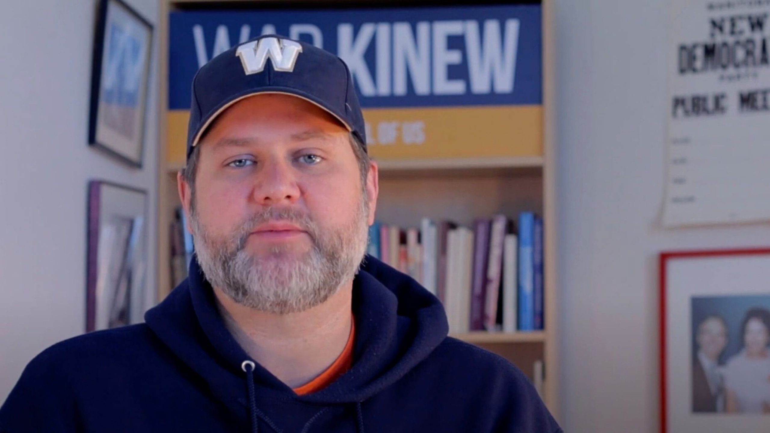 Matt Wiebe wearing a navy blue cap with a "W" logo and a matching hoodie stands indoors, with a bookshelf and framed posters visible in the background.