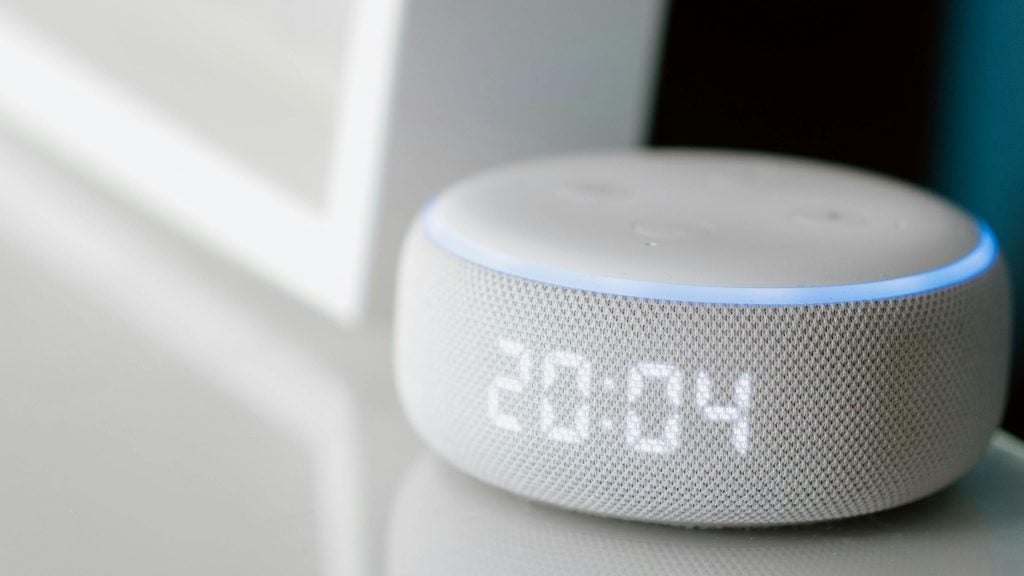 A white smart speaker with a digital clock display showing 20:04, featuring a circular design and a light blue indicator ring on top.