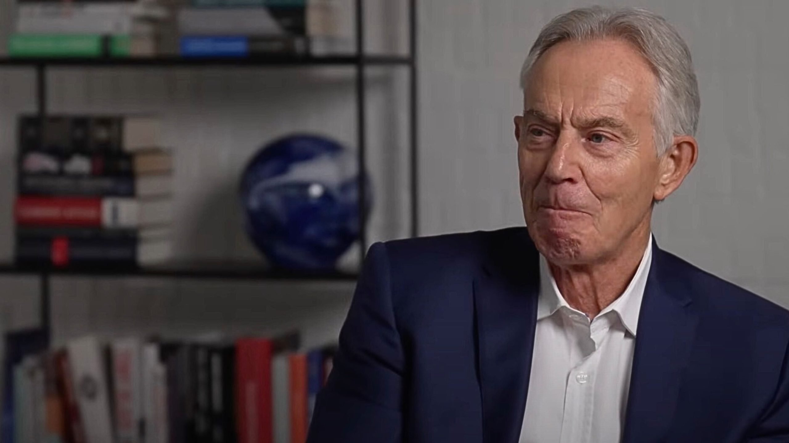 Tony Blair in a suit sitting in front of a bookshelf filled with books and a blue spherical object.