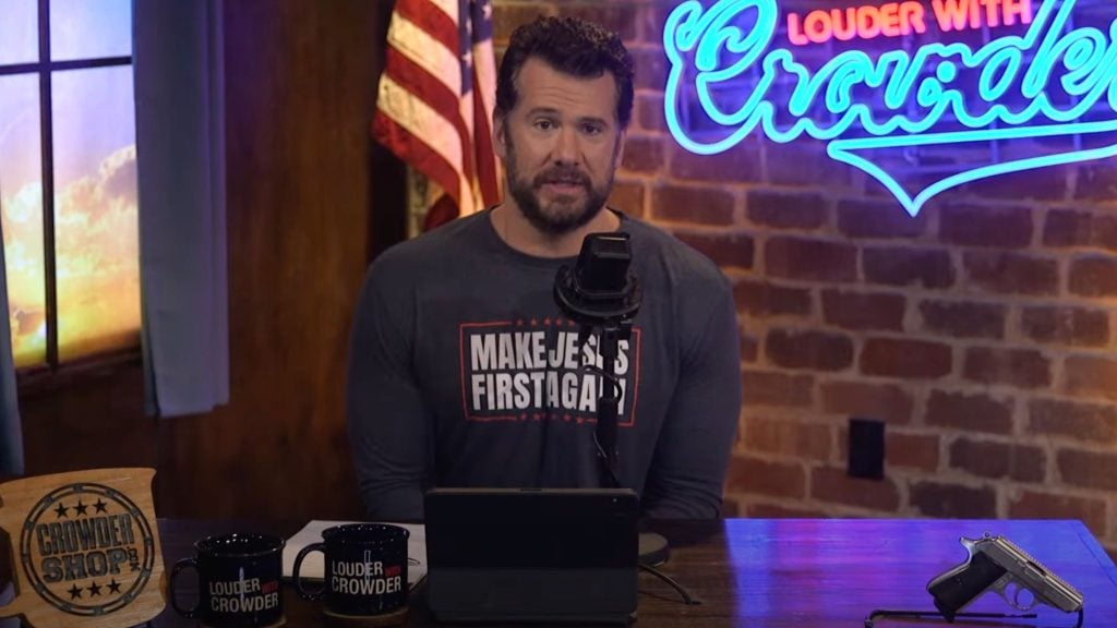 Crowder sits at a podcast or radio studio desk, with an American flag and a neon sign reading "Louder with Crowder" in the background. He is wearing a shirt with the text "Make Jesus First Again". The desk has a microphone, two mugs with the same "Louder with Crowder" logo, a tablet, and a handgun.