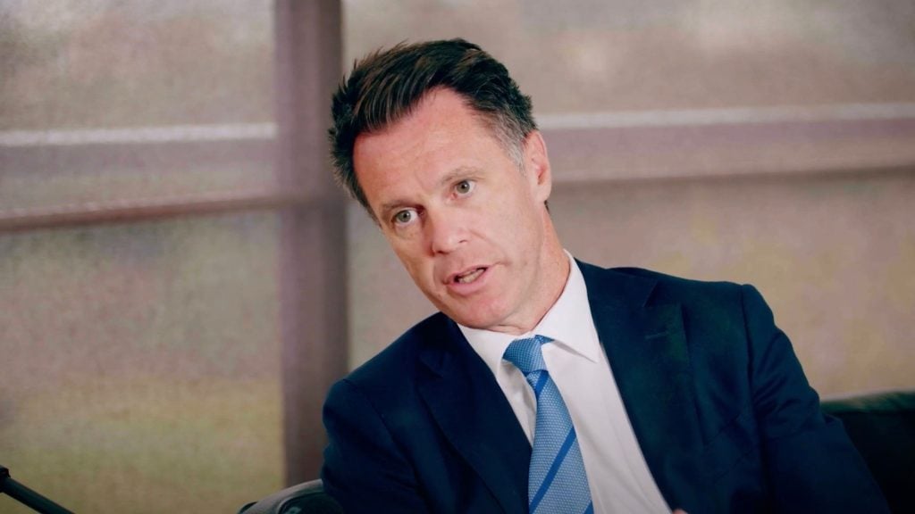 Minns in a suit and tie speaking indoors with a serious expression, sitting in front of a window with blurred outdoor view.