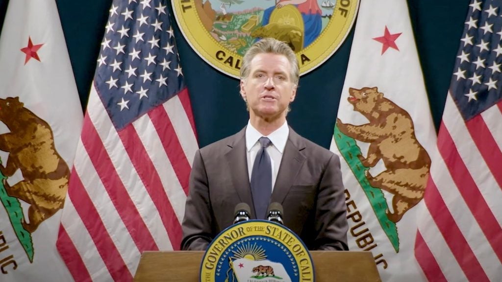 Newsom standing at a podium with the seal of the State of California and surrounded by American and Californian flags.