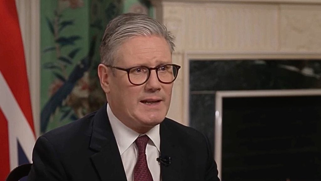 Starmer in a suit and tie speaking indoors, with glasses on, against a background featuring a Union Jack and decorative wallpaper.