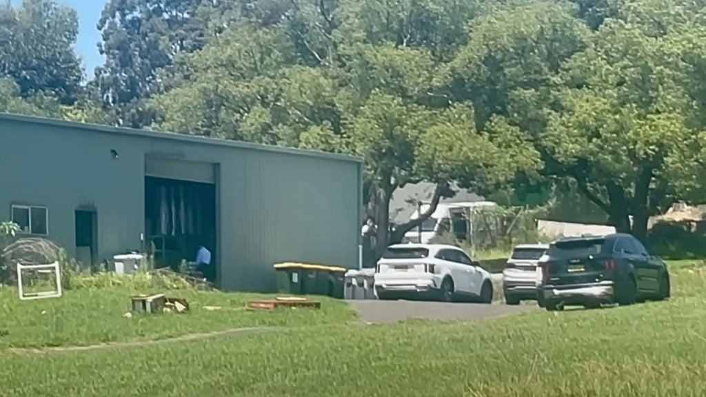 A rural scene with a metal shed featuring an open garage door, several trash bins outside, and two parked cars on the driveway, surrounded by lush trees and grass.