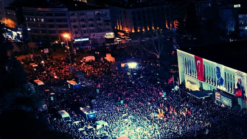 A large crowd gathered in a city square at night, with numerous flags visible, including the Turkish flag. A nearby building displays a large banner with a person’s image and a Turkish flag. The area is brightly lit with streetlights and building lights, and several vehicles are parked amongst the people.