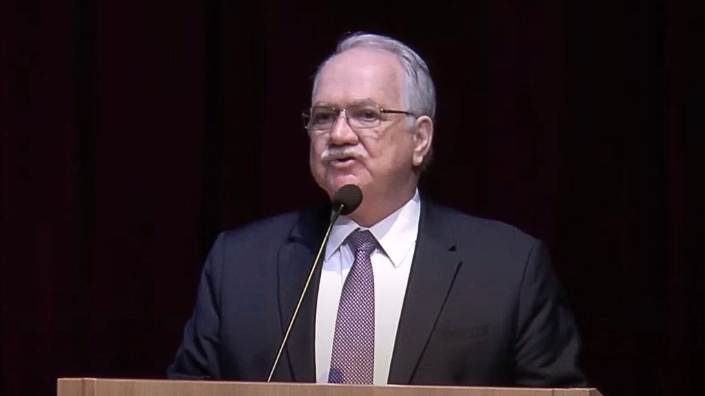 Luiz Edson Fachin with gray hair and glasses speaking at a podium with a microphone, wearing a dark suit, white shirt, and patterned tie against a dark background