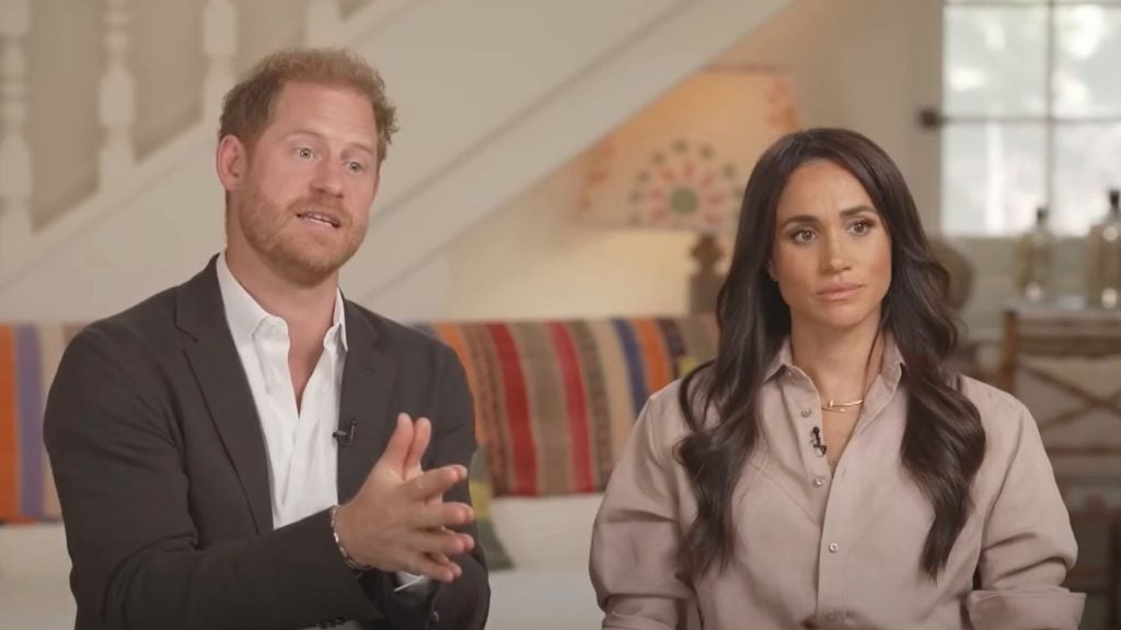 Harry with light brown hair and beard wearing a black suit jacket over a white shirt gestures with his hands while speaking next to Meghan with long dark hair wearing a beige button-up shirt, sitting on a couch with a colorful striped blanket and a patterned lamp in the background.