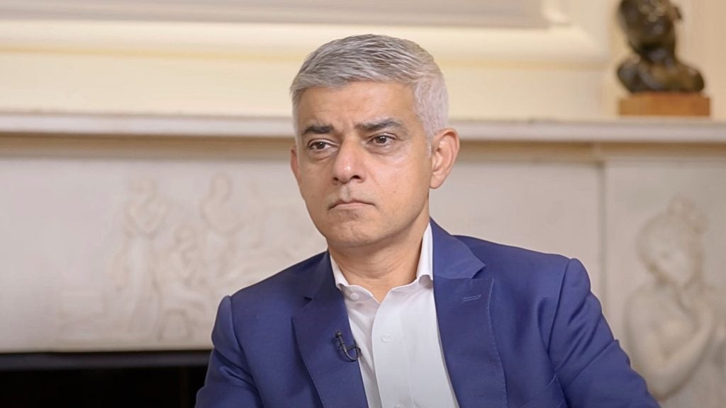 Khan with short gray hair wearing a blue suit and white shirt sitting indoors with a serious expression, in front of a beige wall with decorative carvings and a small statue in the background.