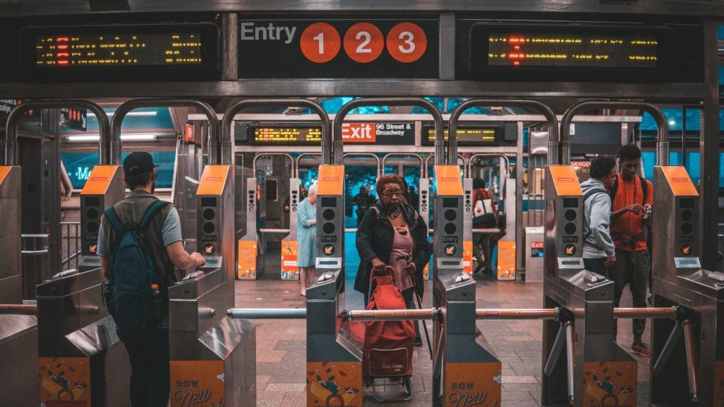 People passing through subway turnstiles with signs indicating entry for lines 1, 2, and 3 and an exit for 96 Street and Broadway in the background.