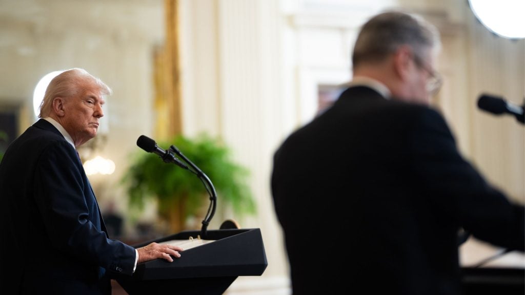 Side view of President Trump with blond hair standing behind a podium with a microphone, looking serious, with Keir Starmer in a suit blurred in the foreground speaking at a microphone in an elegant room with a large mirror and greenery in the background.