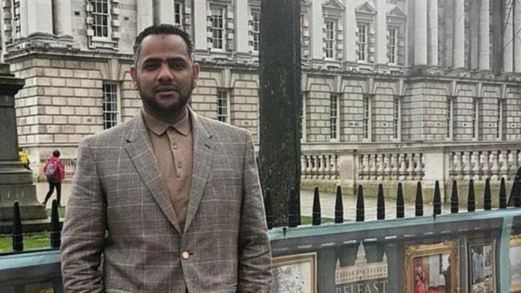 Ahmed al-Doush wearing a checked blazer and beige shirt standing outdoors in front of a historic stone building with tall windows and a decorative fence.