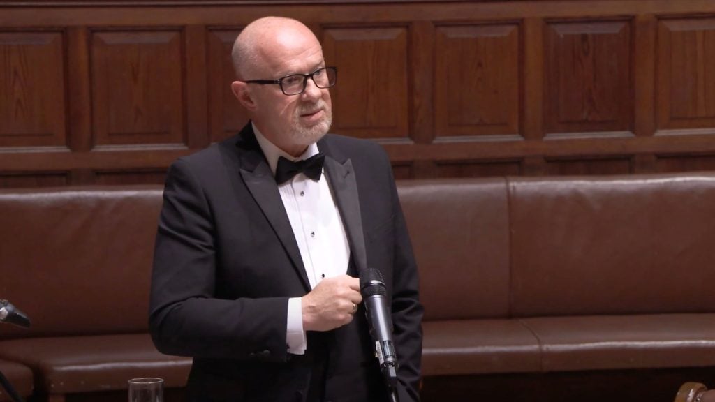 UK's Chief Inspector of Constabulary Andy Cooke with glasses and a beard wearing a black tuxedo and bow tie, standing and speaking in front of a microphone with a wooden panel and brown leather bench background.