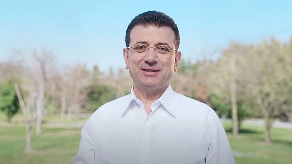 Istanbul Mayor Ekrem Imamoglu wearing glasses and a white shirt standing outdoors with trees and grass in the background under a clear blue sky.