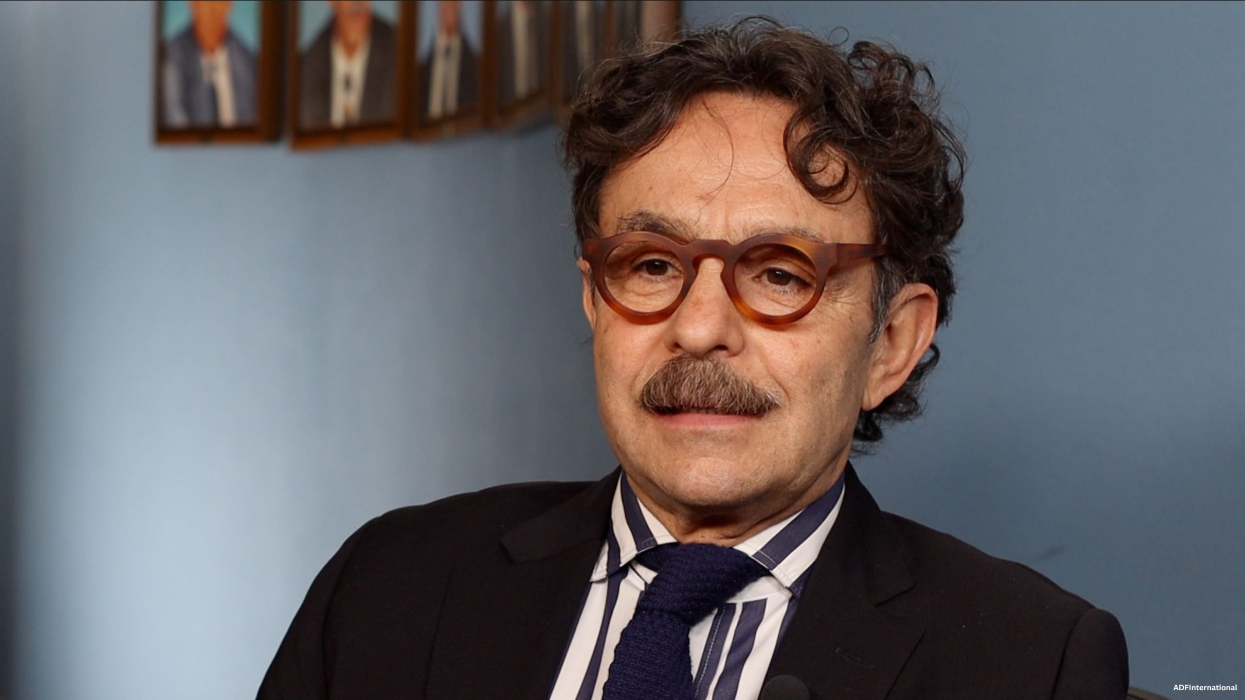 Middle-aged man with curly dark hair, a mustache, and tortoiseshell glasses wearing a black suit, striped shirt, and dark knitted tie sitting against a blue wall with framed photos in the background.