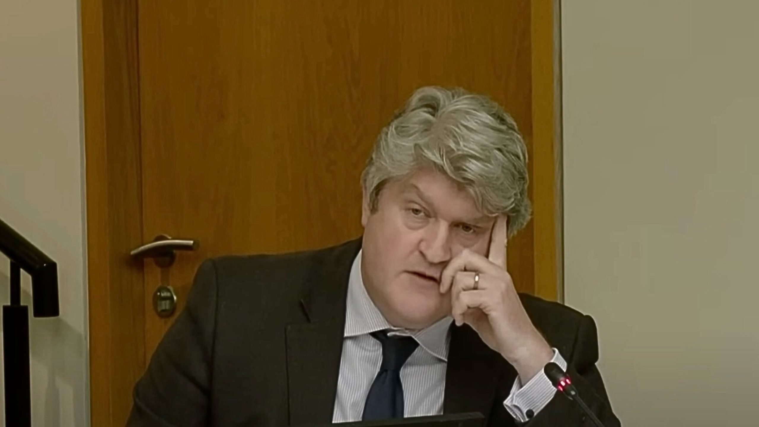 Michael McNamara with gray hair wearing a black suit, white shirt, and dark tie, resting his head on his hand with a contemplative expression, seated in front of a wooden door with a microphone nearby.
