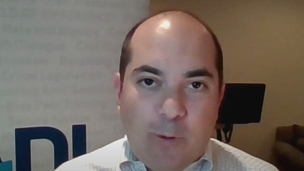 ADL's Midwest Regional Director David Goldenberg wearing a white shirt with a patterned collar, speaking indoors with a blurred background featuring a dark monitor and letters partially visible on the left.