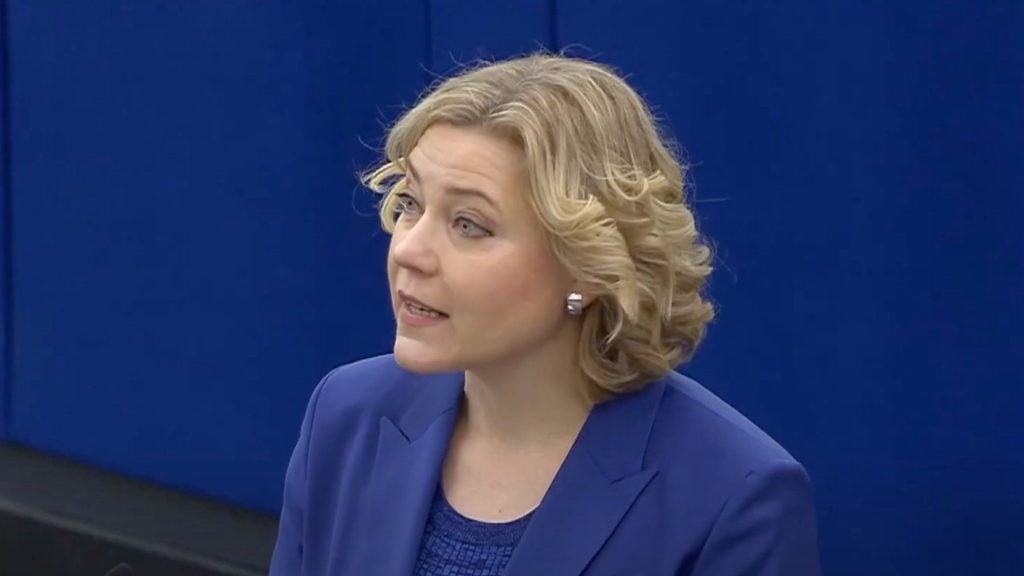 Henna Virkkunen with short curly blonde hair wearing a blue blazer and top speaking against a blue background.