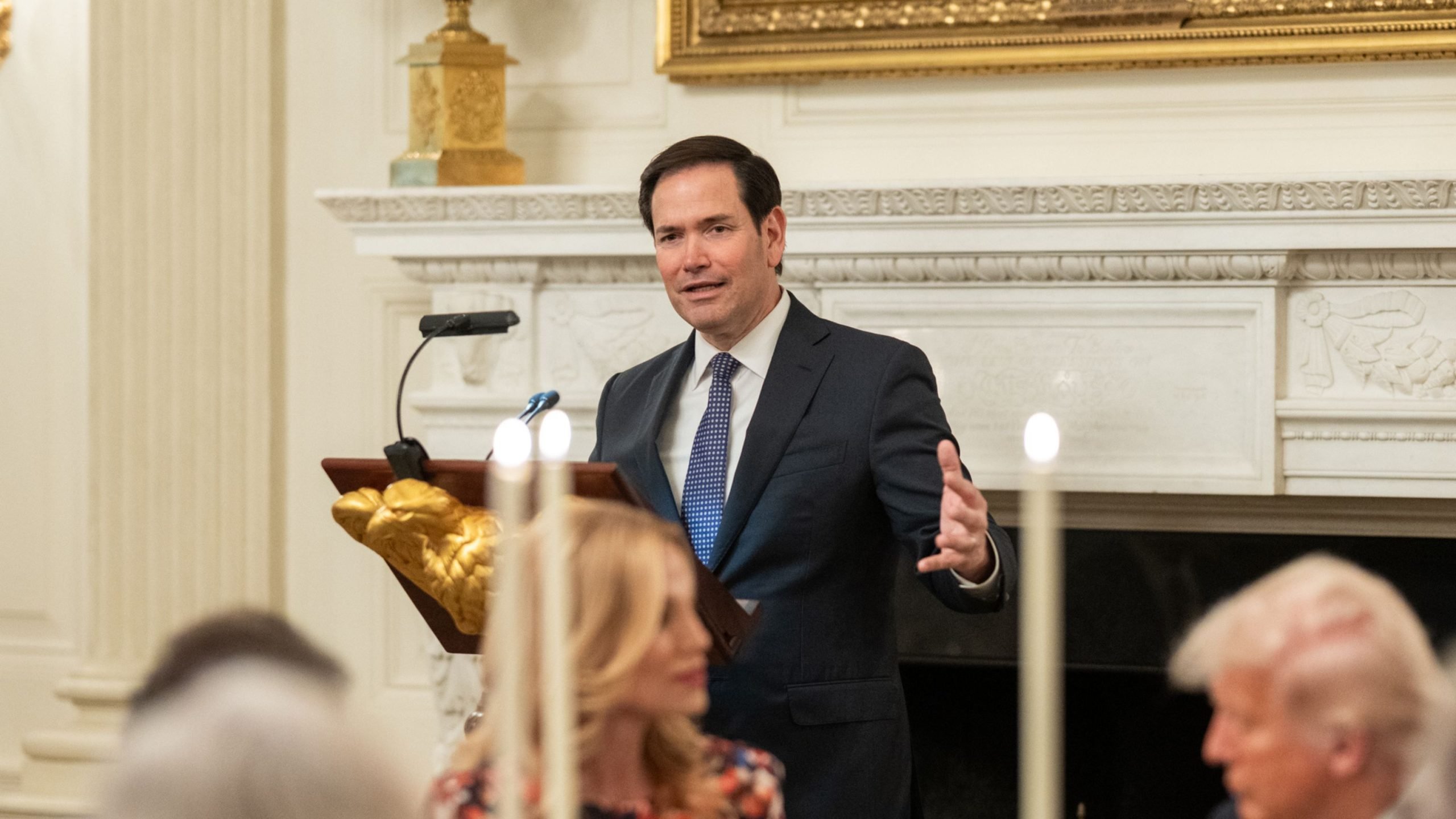 Marco Rubio in a suit speaking at a podium with a microphone in a formal room, with a blurred woman and an older man with light hair in the foreground, and a decorative white fireplace in the background.