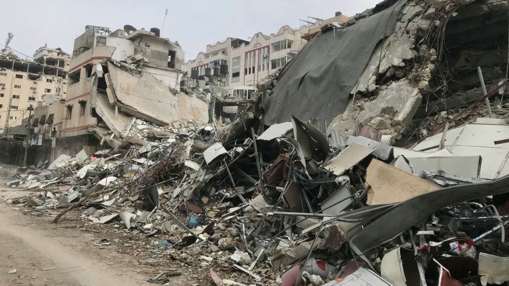 Street view showing extensive rubble and debris from collapsed and severely damaged buildings under a gray sky.