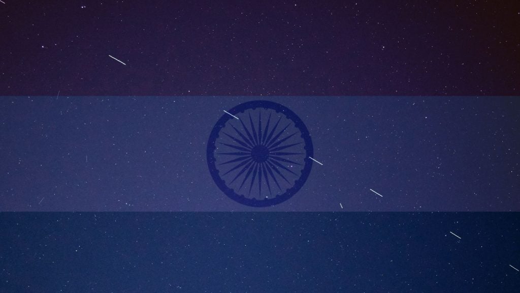 Night sky with numerous stars and streaks of light overlaid with a transparent Indian flag featuring the Ashoka Chakra in the center.