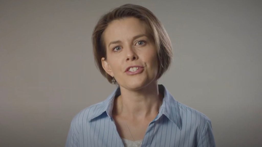 Woman with short brown hair wearing a light blue striped shirt speaking against a plain gray background.