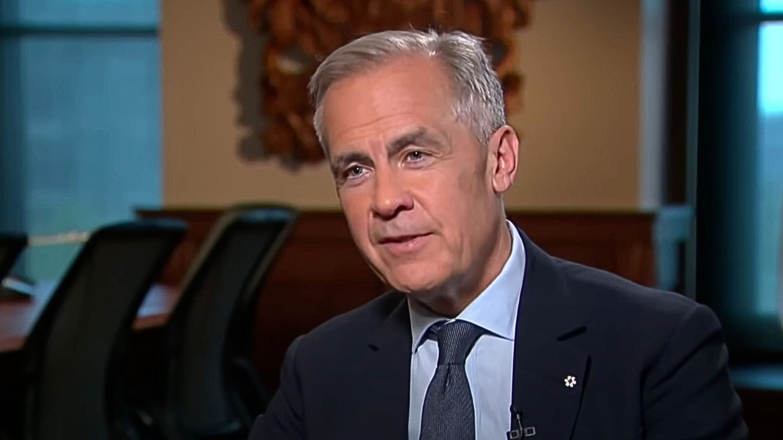 Carney with gray hair wearing a dark suit and blue tie, seated indoors with blurred office chairs and a window in the background.