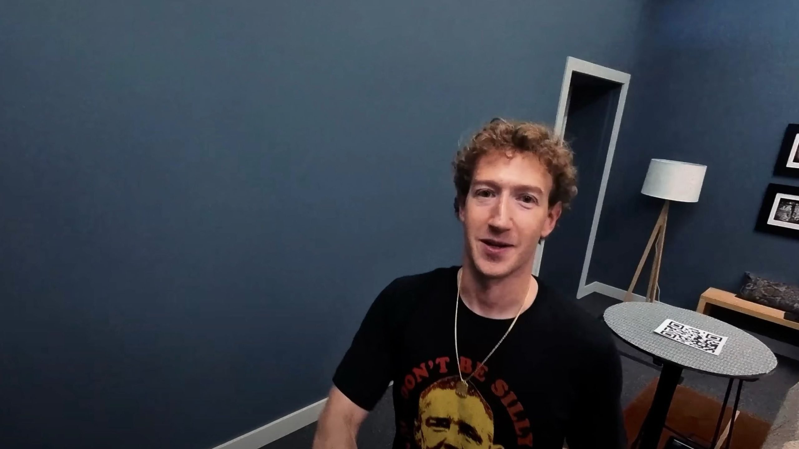 Zuckerberg with curly hair wearing a black t-shirt with a graphic and text, standing in a modern room with blue walls, a round table with a QR code on it, a floor lamp, and framed pictures on the wall.