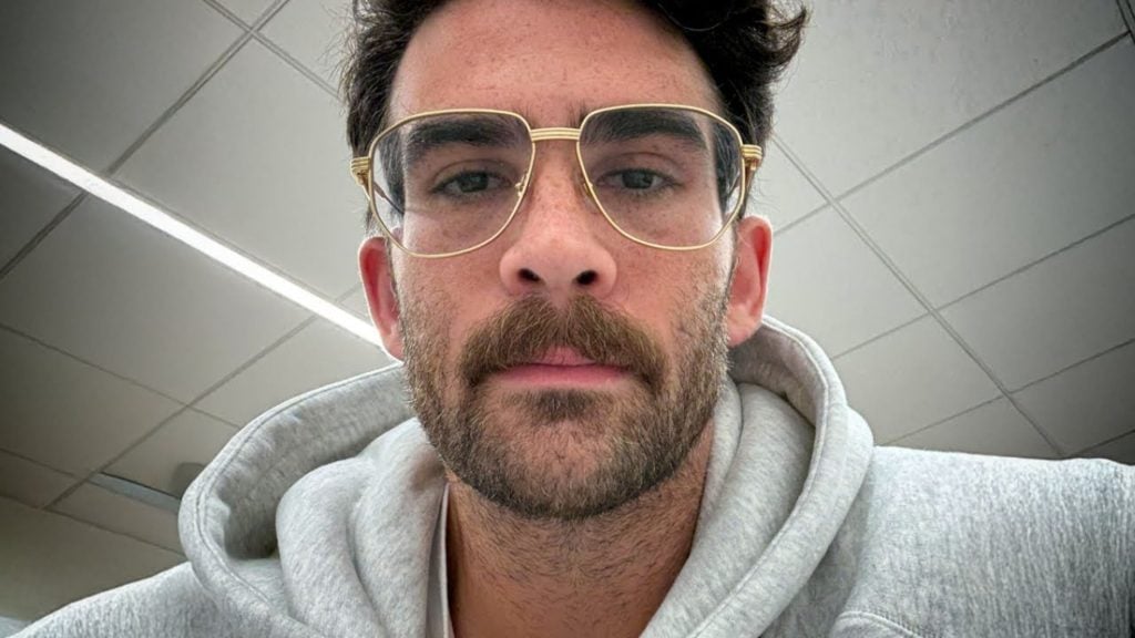 Piker with dark hair, a mustache and beard, wearing a light gray hoodie and gold-rimmed glasses, with a ceiling of square tiles in the background.