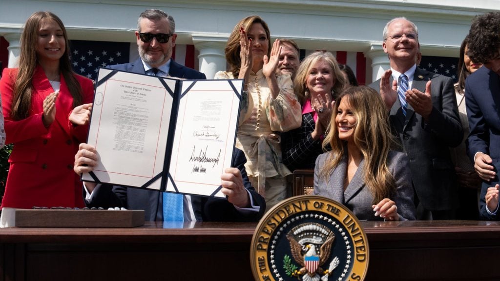 Trump seated at a desk holding up a signed document in front of his face while smiling people stand behind him applauding outside a government building with American flags and presidential seal on the desk.