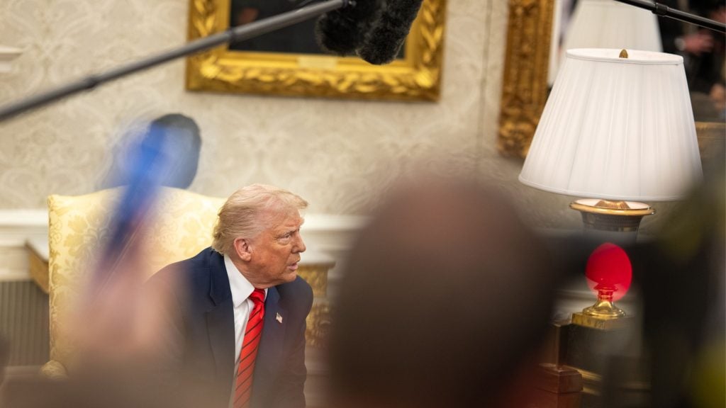Trump with blonde hair wearing a dark suit and red striped tie sitting in a light yellow patterned chair in an ornate room with a white lamp and framed painting in the background, partially obscured by blurred microphones and heads in the foreground.