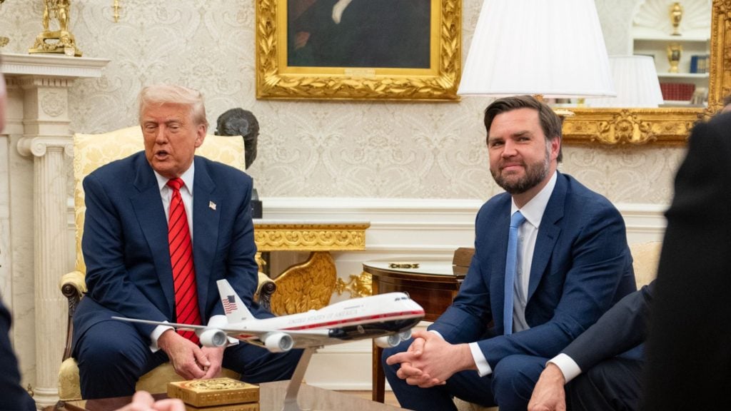 Trump and Vance in suits seated in a formal room with ornate decor, a small model airplane on the table between them, and a large framed portrait and lamp in the background.