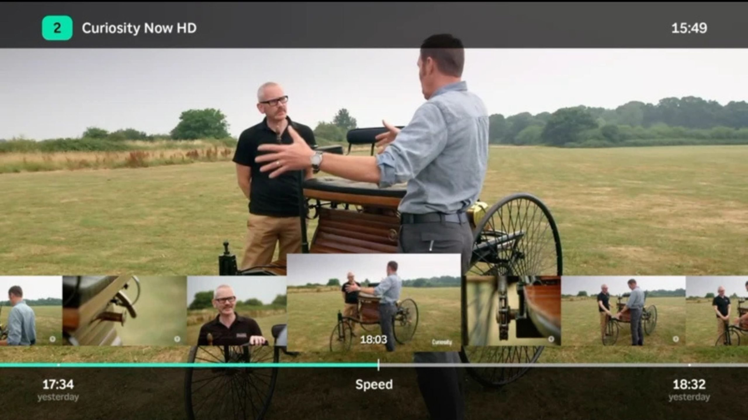 Two men talking outside in a grassy field near a vintage three-wheeled vehicle, with a video playback interface showing thumbnails and a timeline bar overlaid on the scene.