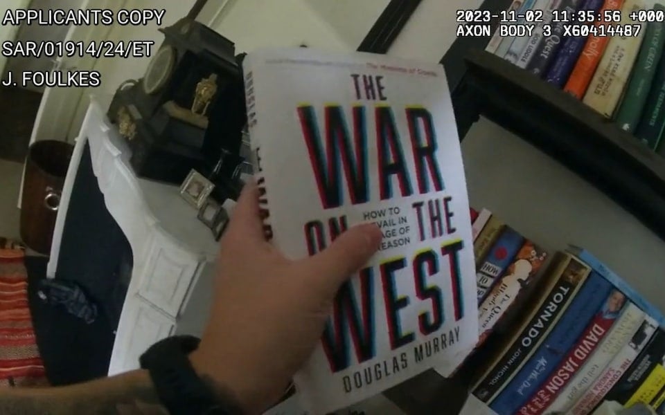Hand holding the book titled 'The War on the West' by Douglas Murray in front of a bookshelf filled with various books and a white fireplace mantle with a clock and picture frames on top.