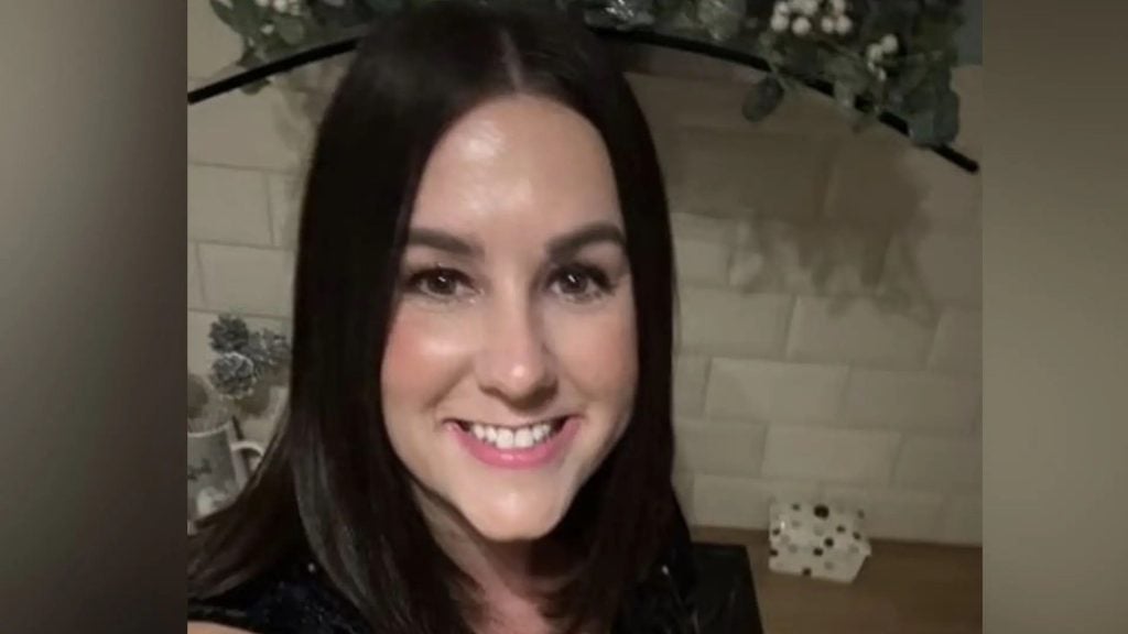 Close-up of a smiling Connolly with straight dark hair, wearing a black top, standing indoors in front of a white tiled wall and a shelf with decorative items and greenery.
