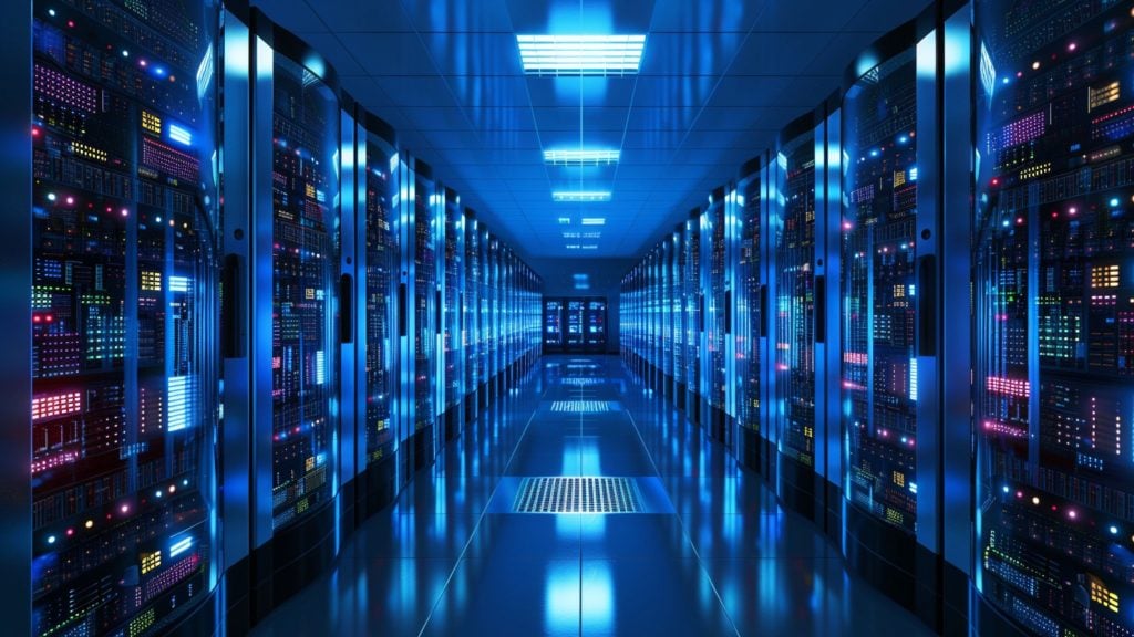 Long corridor in a data center lined with rows of brightly lit server racks emitting blue, red, yellow, and green indicator lights under fluorescent ceiling lights.