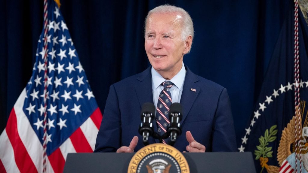 Joe Biden standing at a podium with the presidential seal, speaking with two microphones, with American flags in the background.