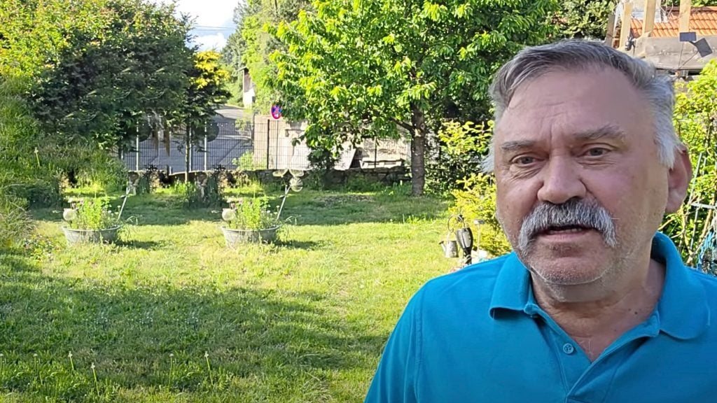 Stefan Niehoff with gray hair and mustache wearing a blue polo shirt standing in a sunny garden with green grass, potted plants, and trees in the background.